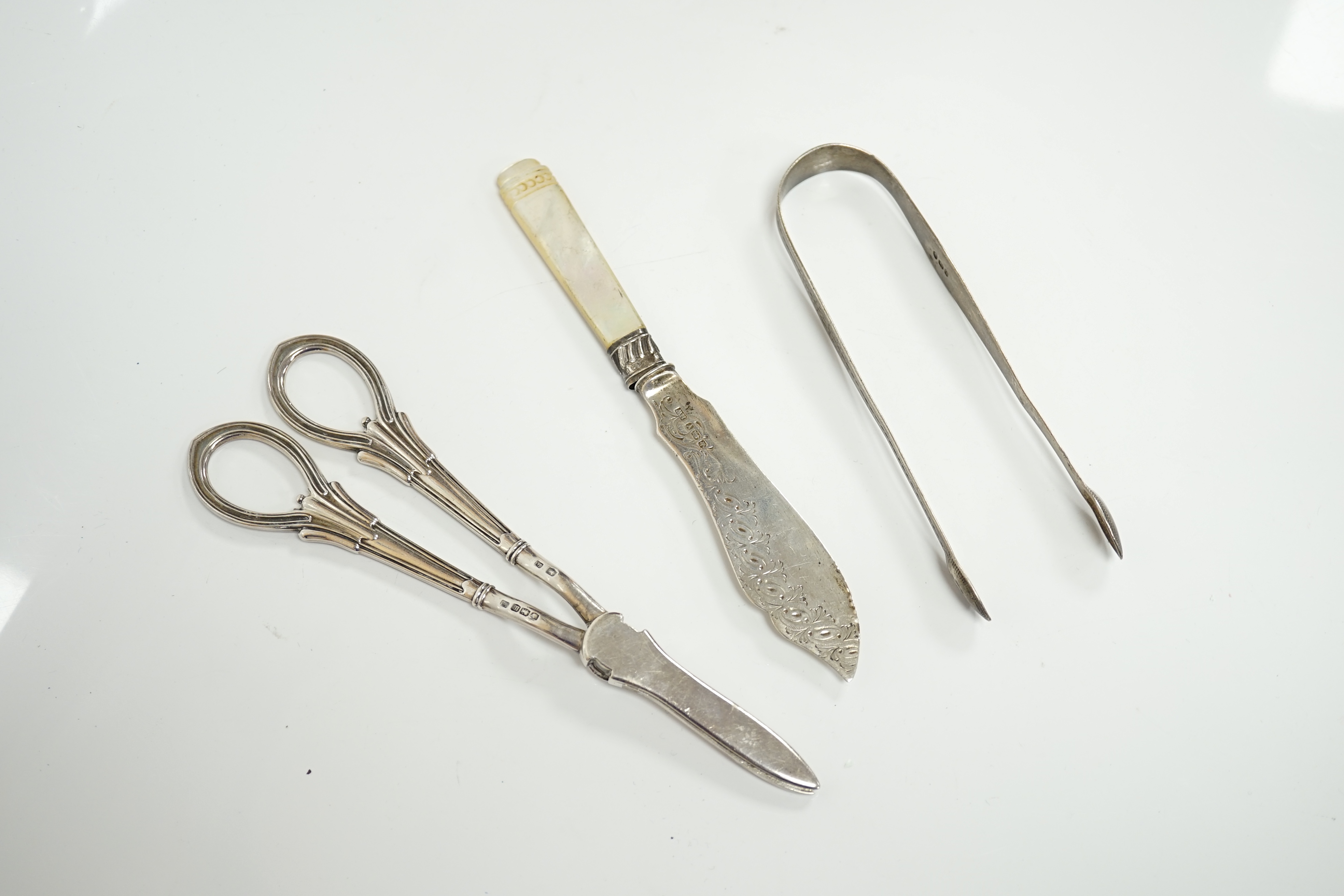 A pair of George V silver grape scissors, Sheffield, 1929, 18.6cm, together with a mother of pearl handled silver butter knife ad a pair of Georgian silver sugar tongs by Peter & Ann Bateman. Condition - poor to fair